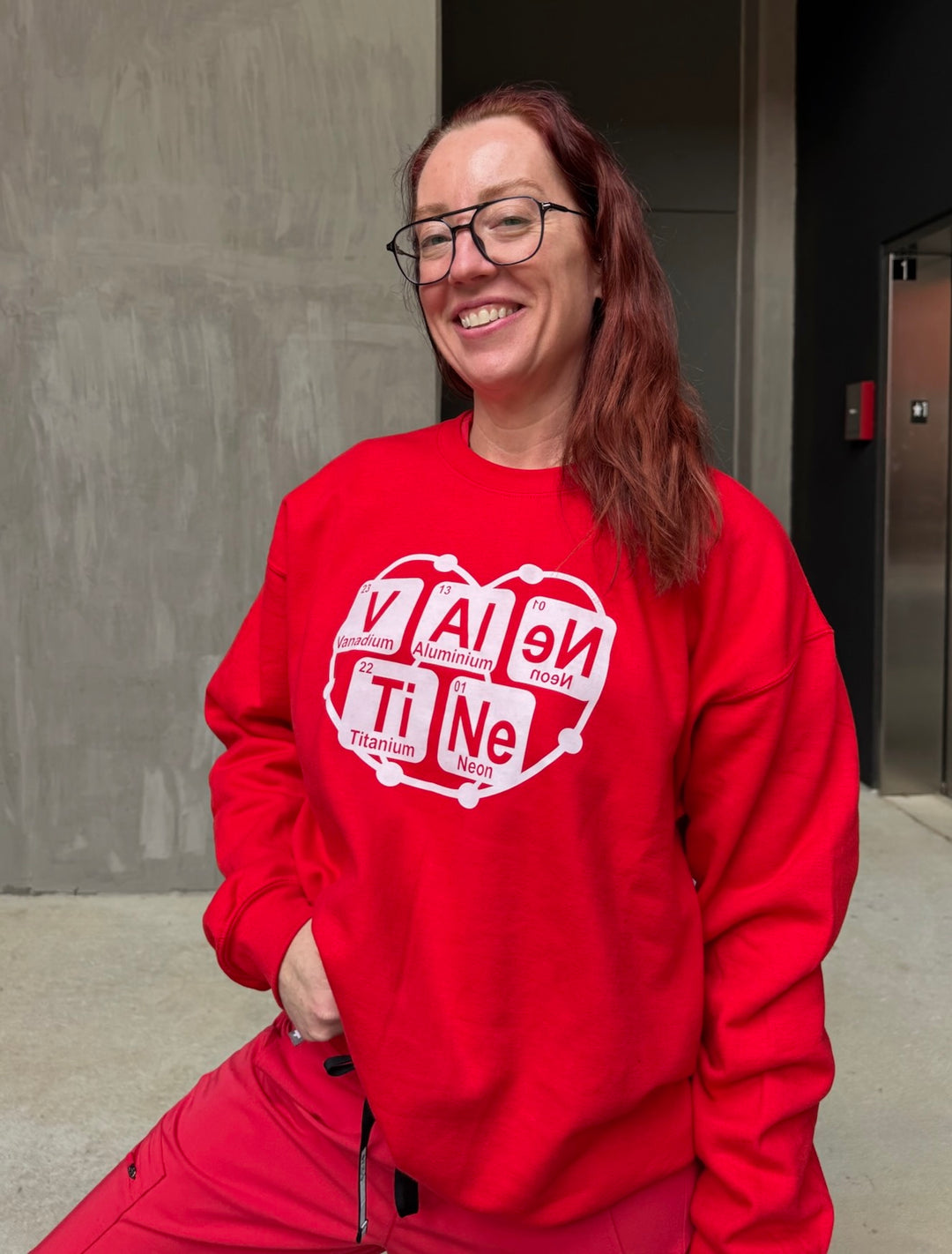 Ladyspinedoc wearing a red sweatshirt with a white graphic Valentine periodic table design, standing against a plain wall.