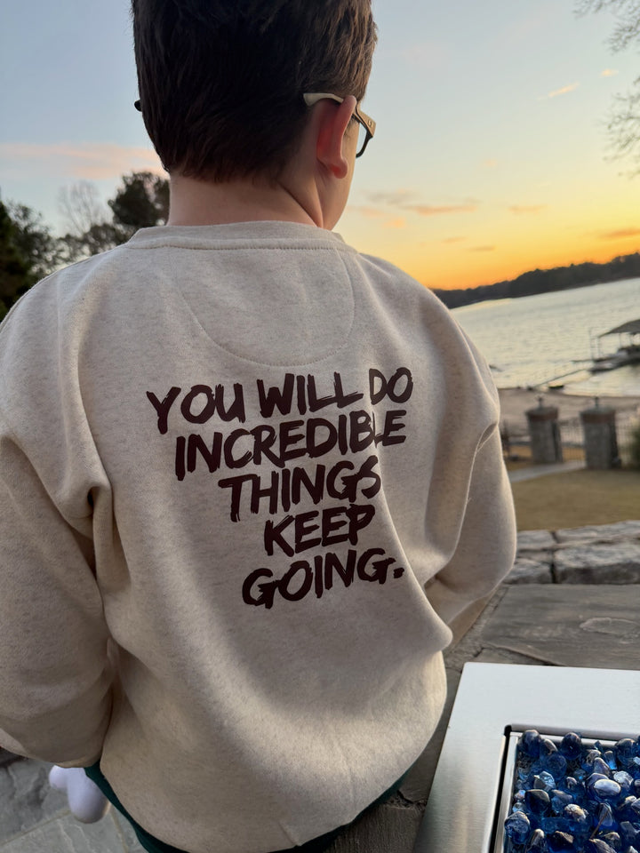 Youth wearing the Incredible Crewneck Youth Sweatshirt - Kids Natural Heather Shirt with Motivational "YOU WILL DO INCREDIBLE THINGS. KEEP GOING." motto on the back, standing by a lake at sunset.