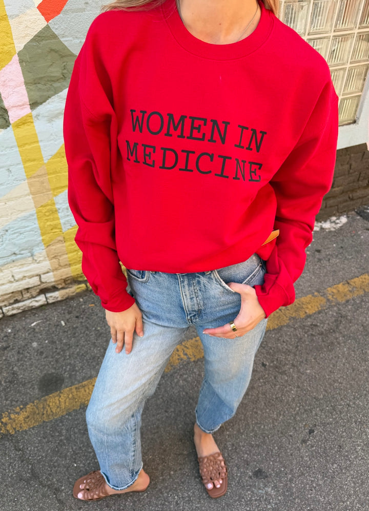 Person wearing a red sweatshirt with black 'WOMEN IN MEDICINE' text, standing on a street.