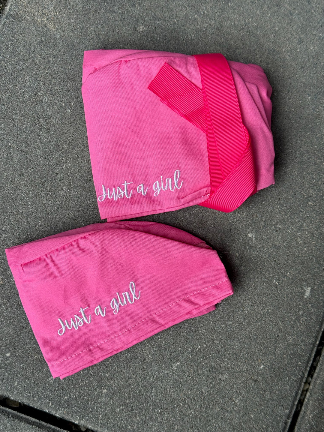 Two pink pairs of scrub hats with 'Just a girl' embroidered text on a gray surface