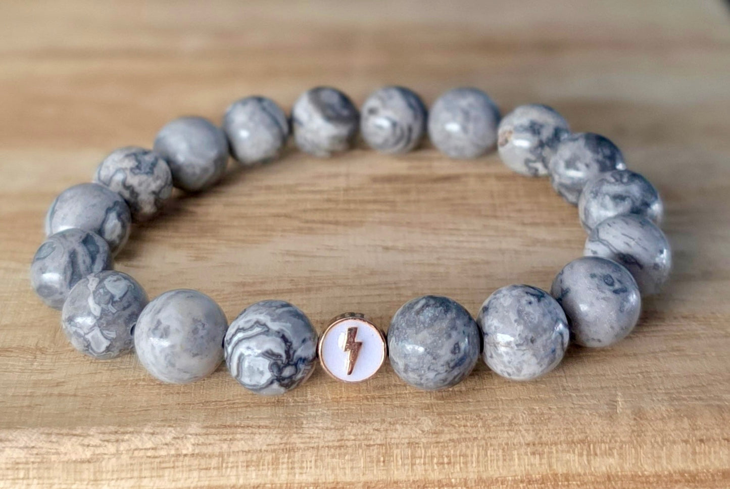 Close up view of Glioblastoma Awareness Bracelet made of gray Map Stone Jasper beads with one white enamel bead with gold lightning bolt.