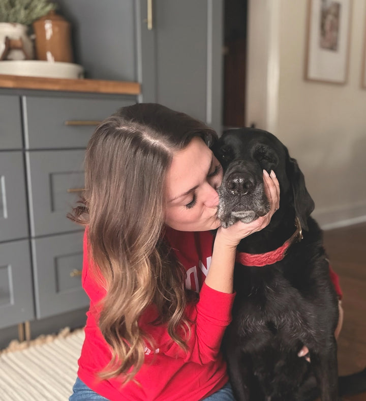 Dr Molly in a red shirt kissing a black dog on the head in a home setting.