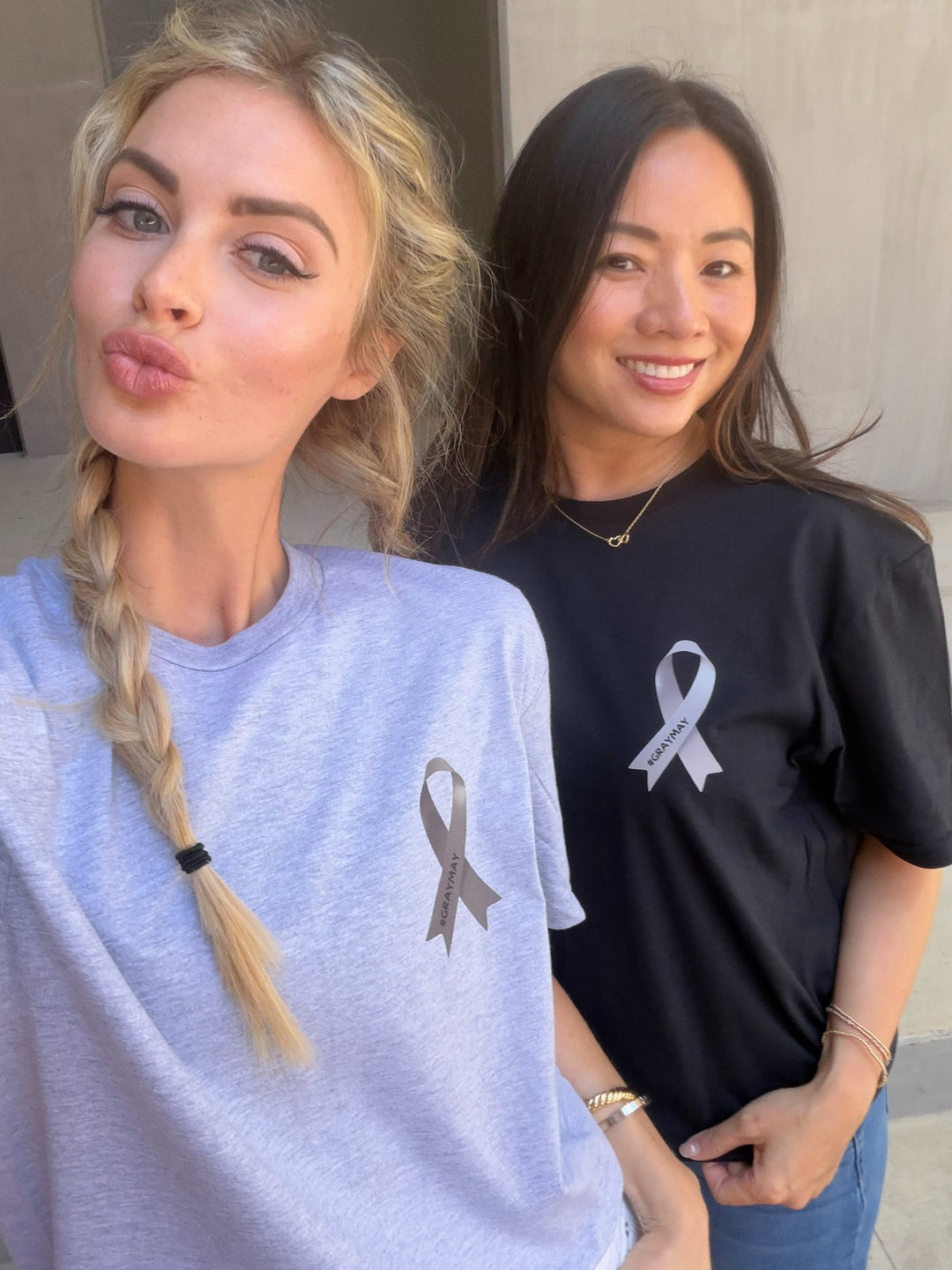 Two women wearing shirts with gray ribbons, for brain tumor awareness month campaign. The ribbon has #graymay on it.