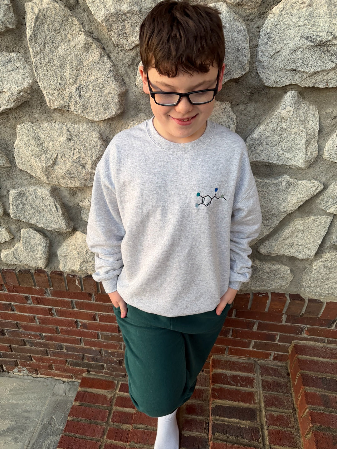 Child against a stone wall wearing a gray sweatshirt with an embroidered Epinephrine (Adrenaline) Molecule Design.