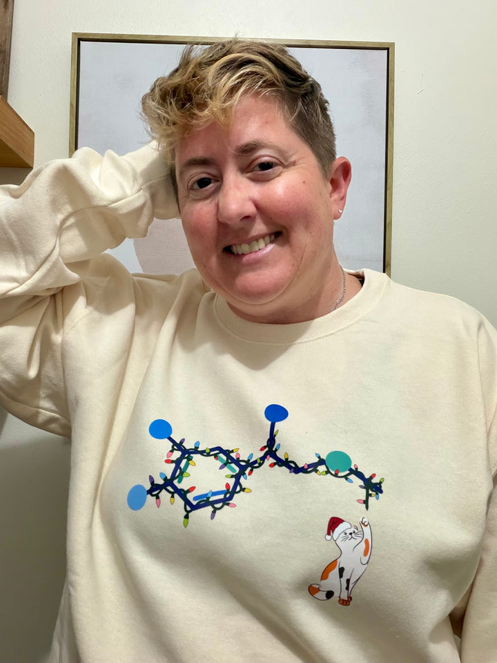 Dr. Beachgem wearing a cream-colored sweatshirt with a Christmas-themed adrenaline cat design, standing indoors.