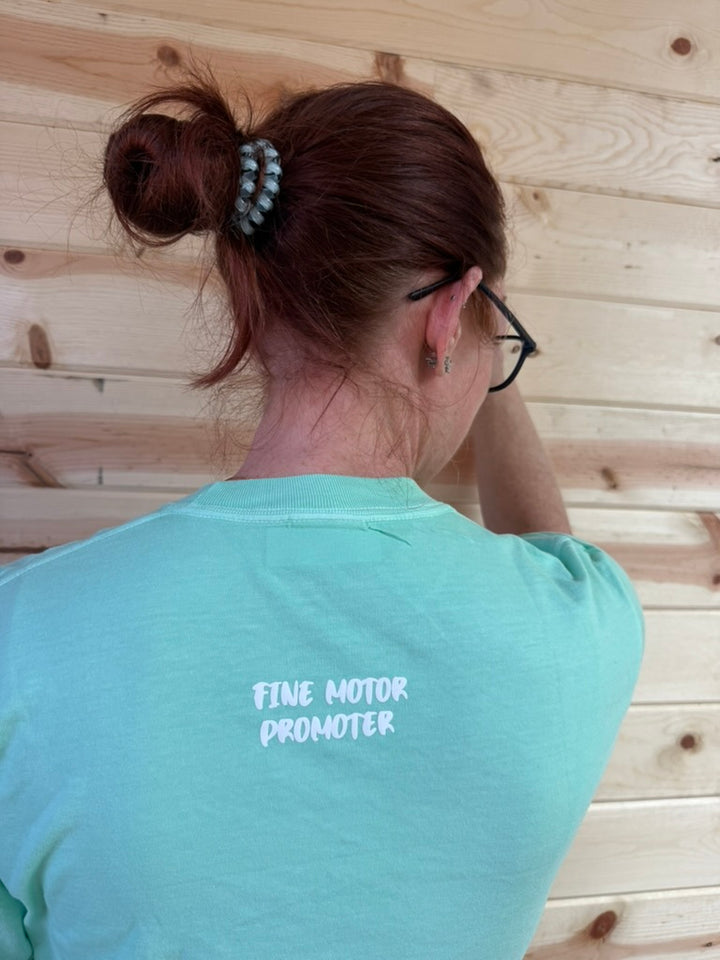 Close-up of the back of a light green T-shirt with "FINE MOTOR PROMOTER" printed in white, worn by Ladyspinedoc (Dr. Betsy Grunch) with her hair up in a bun, standing in front of a wood-paneled wall.
