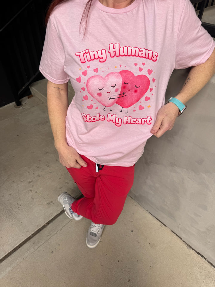 Person wearing a pink t-shirt with heart design and Tiny Humans Stoel My Heart text, standing on a concrete floor.