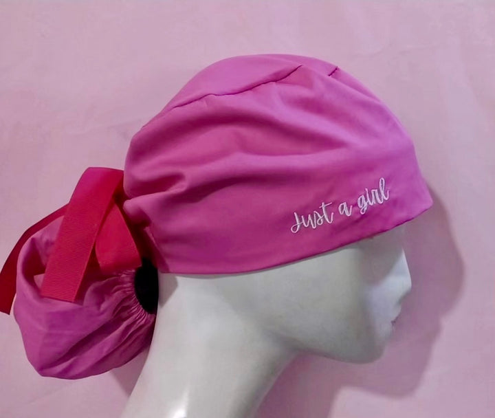 Pink pony tail scrub cap with white embroidered 'Just a girl' text on a mannequin head against a pink background
