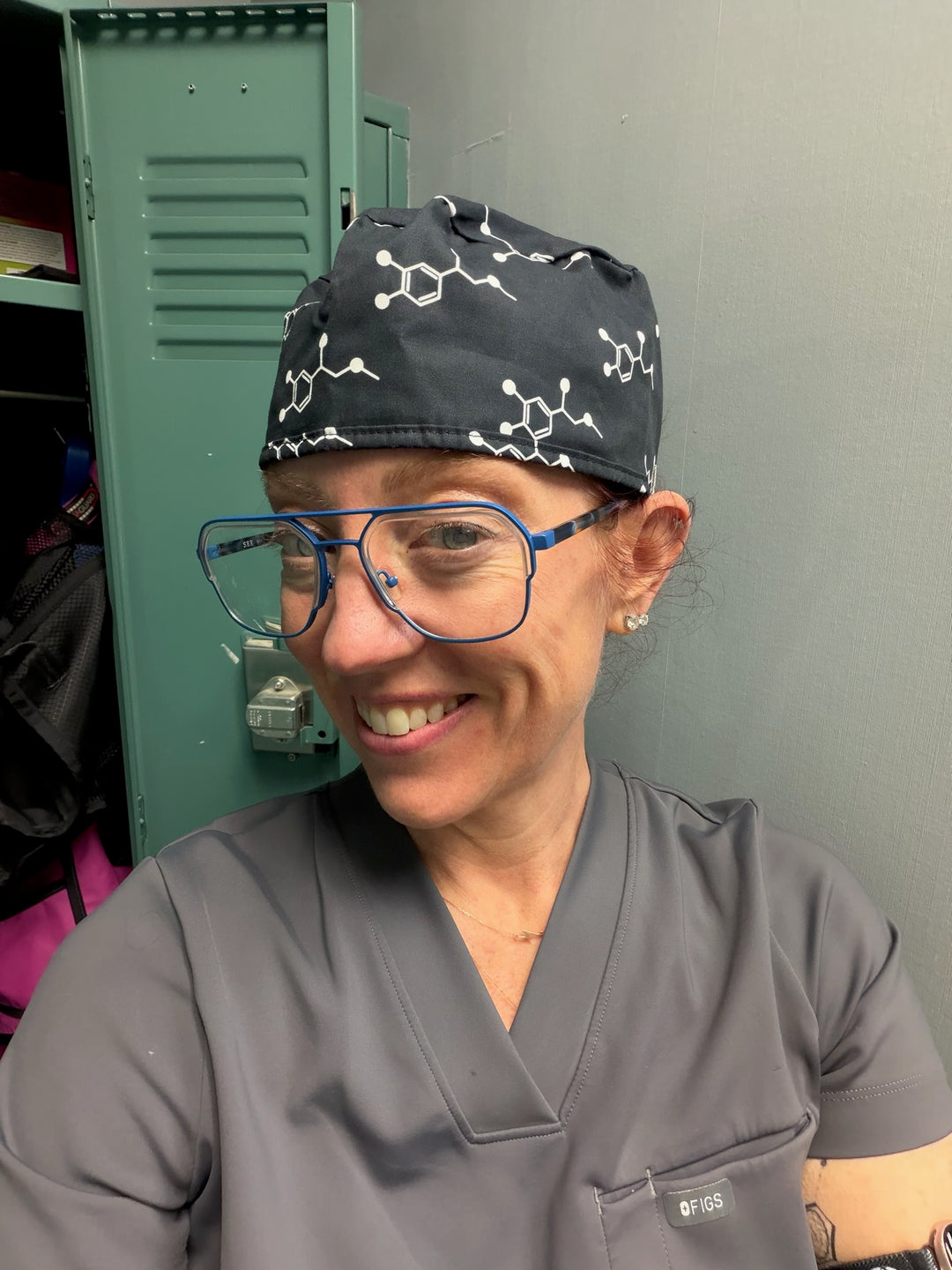 Ladyspinedoc wearing a scrub cap with molecular designs and glasses, standing in front of a green locker.