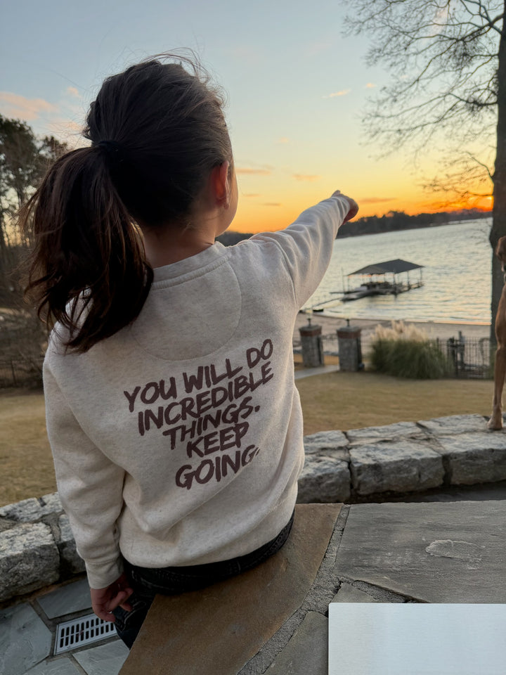Young girl wearing the Incredible Crewneck Youth Sweatshirt - Kids Natural Heather Shirt with Motivational "YOU WILL DO INCREDIBLE THINGS. KEEP GOING." motto on the back, pointing towards a sunset over a lake.