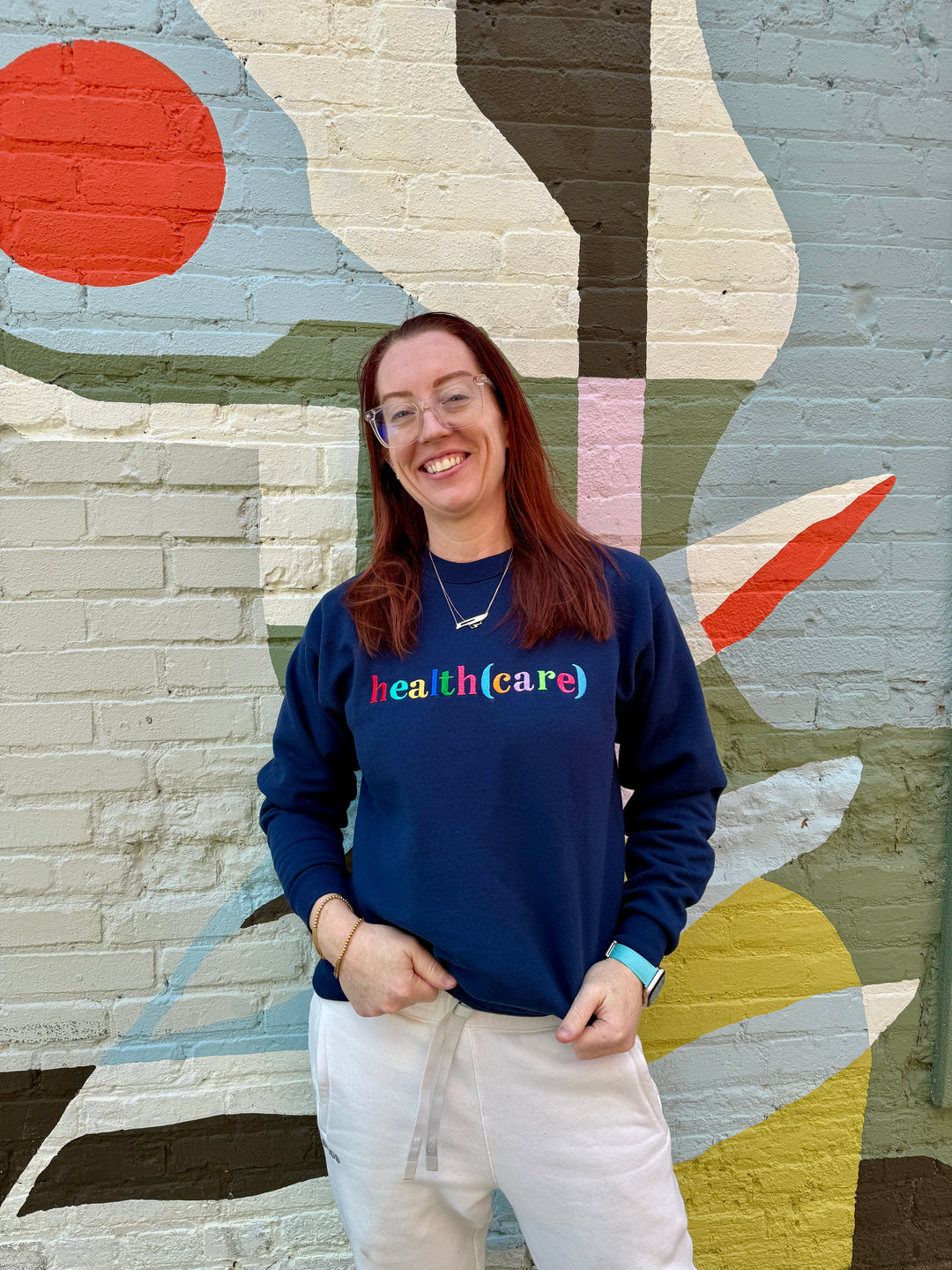 Ladyspinedoc wearing a blue sweatshirt with 'health(care)' text in front of a colorful mural.