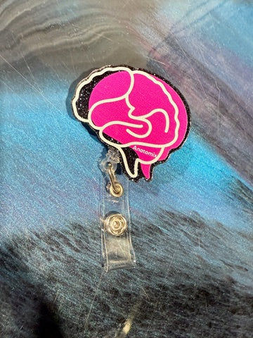 Pink brain-shaped badge holder with the word "Anatomix" at the bottom and laying on a blue and gray background