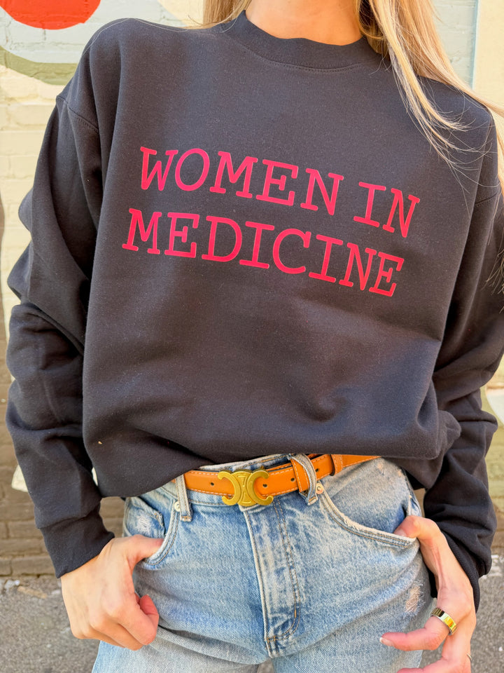 Person wearing a black sweatshirt with 'WOMEN IN MEDICINE' in red text, standing outdoors.