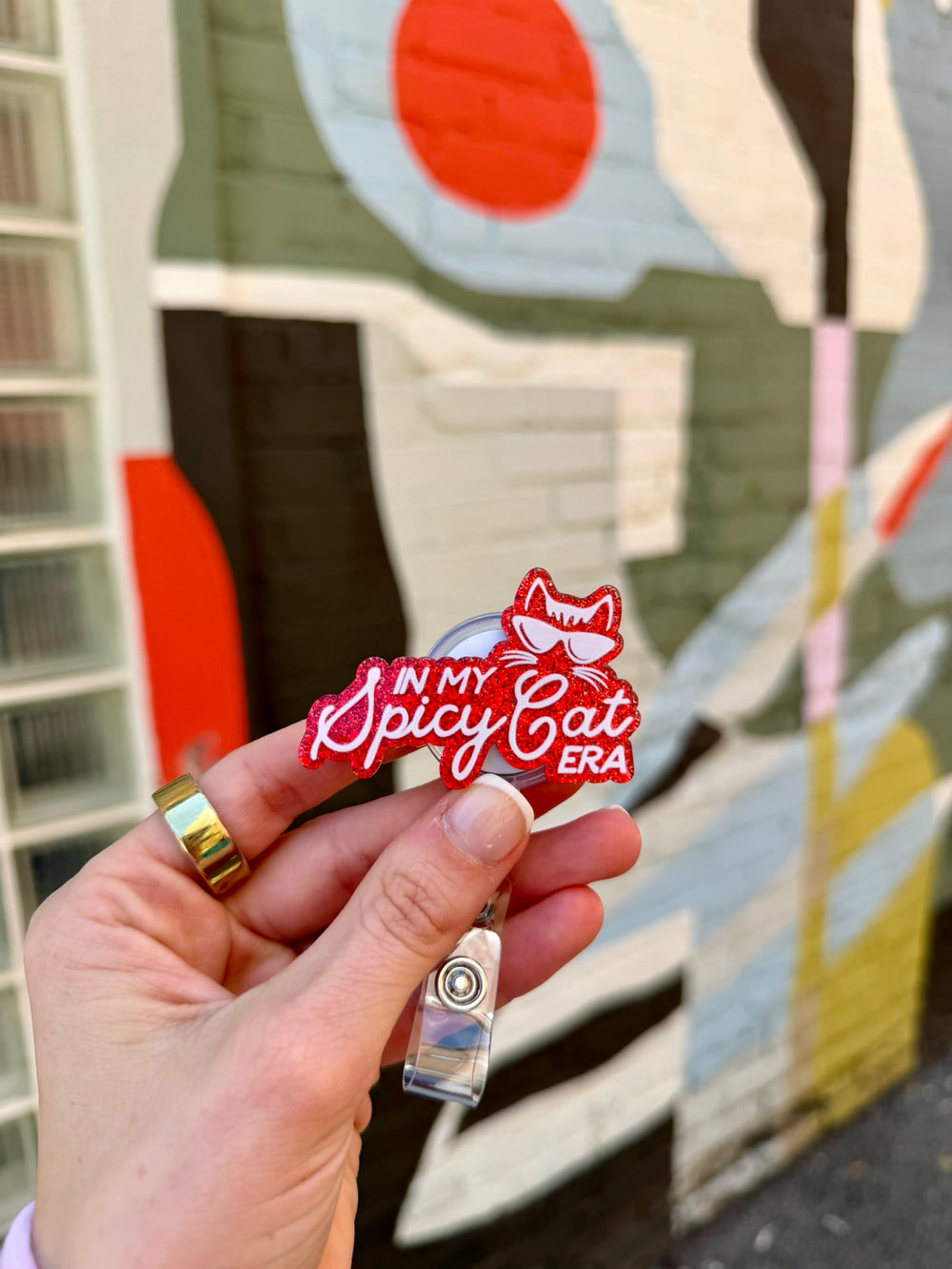 Hand holding a red and white badge reel with In My Spicy Cat Era text against a colorful mural background