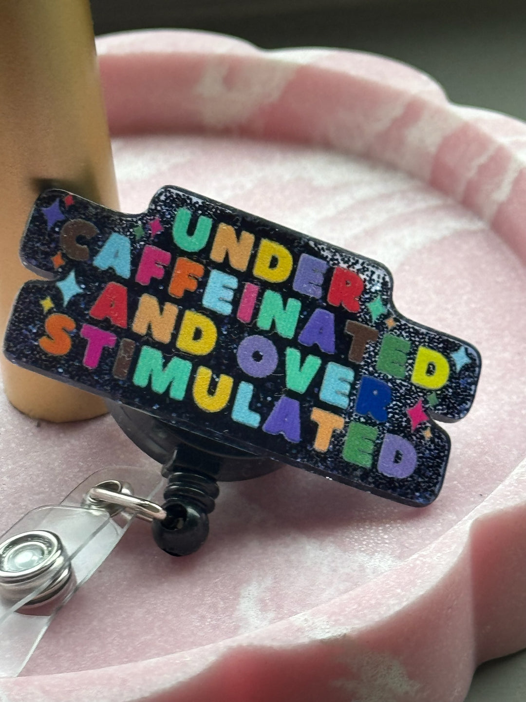 Retractable badge reel with colorful lettering that reads “Under Caffeinated and Over Stimulated” on a sparkly blue and black glitter background and displayed in a pink scallop tray.