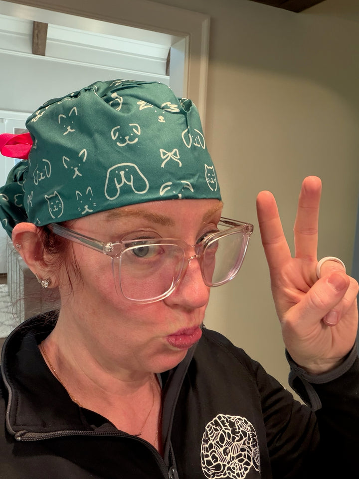Ladyspinedoc wearing a green scrub cap with animal patterns, clear glasses, and a black jacket with a logo, making a peace sign.