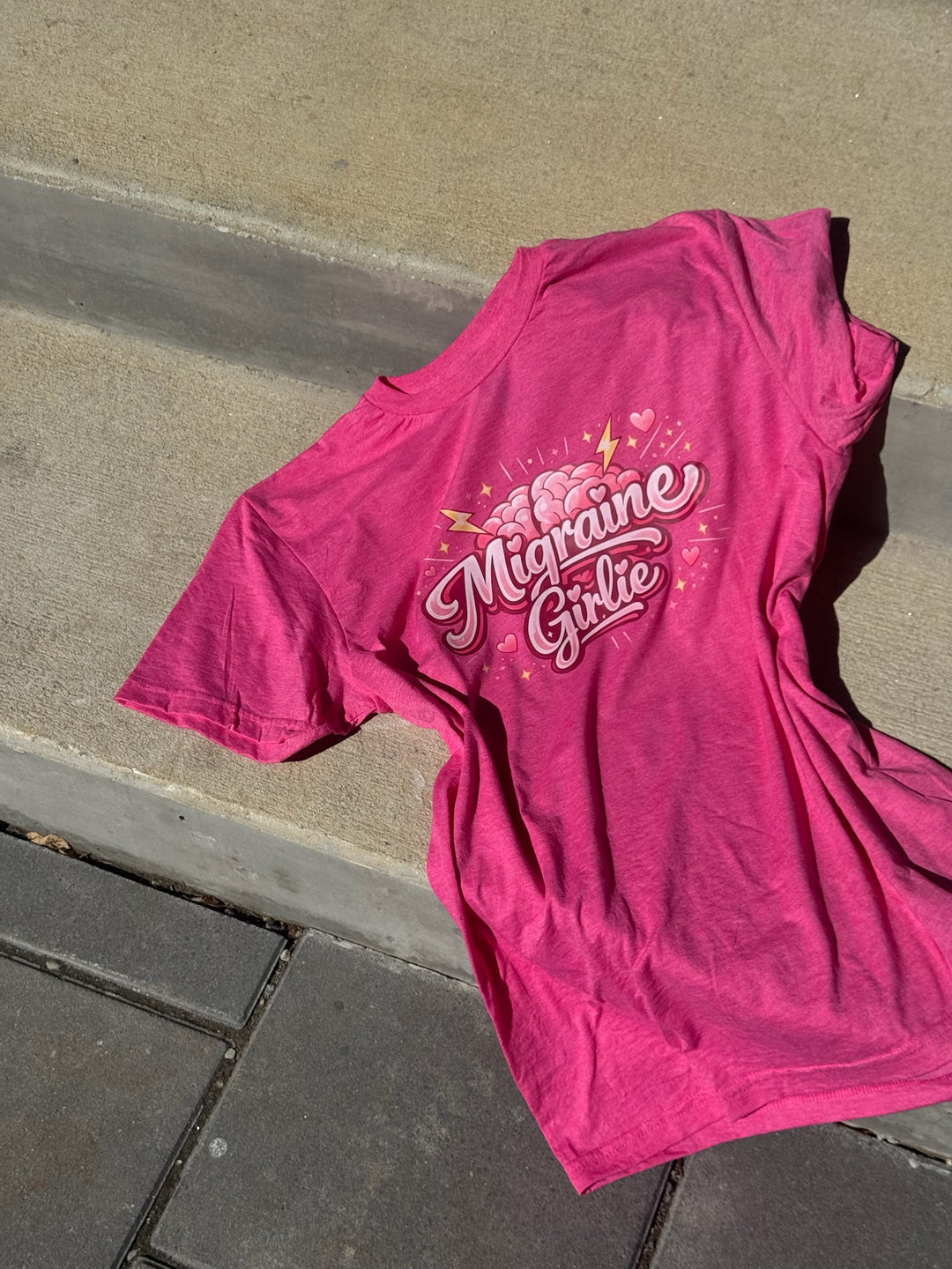 Pink t-shirt with 'Migraine Girlie' text on a concrete surface