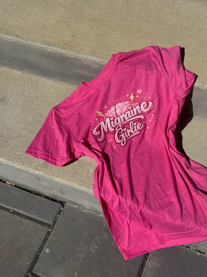 Pink t-shirt with 'Migraine Girlie' text on a concrete surface