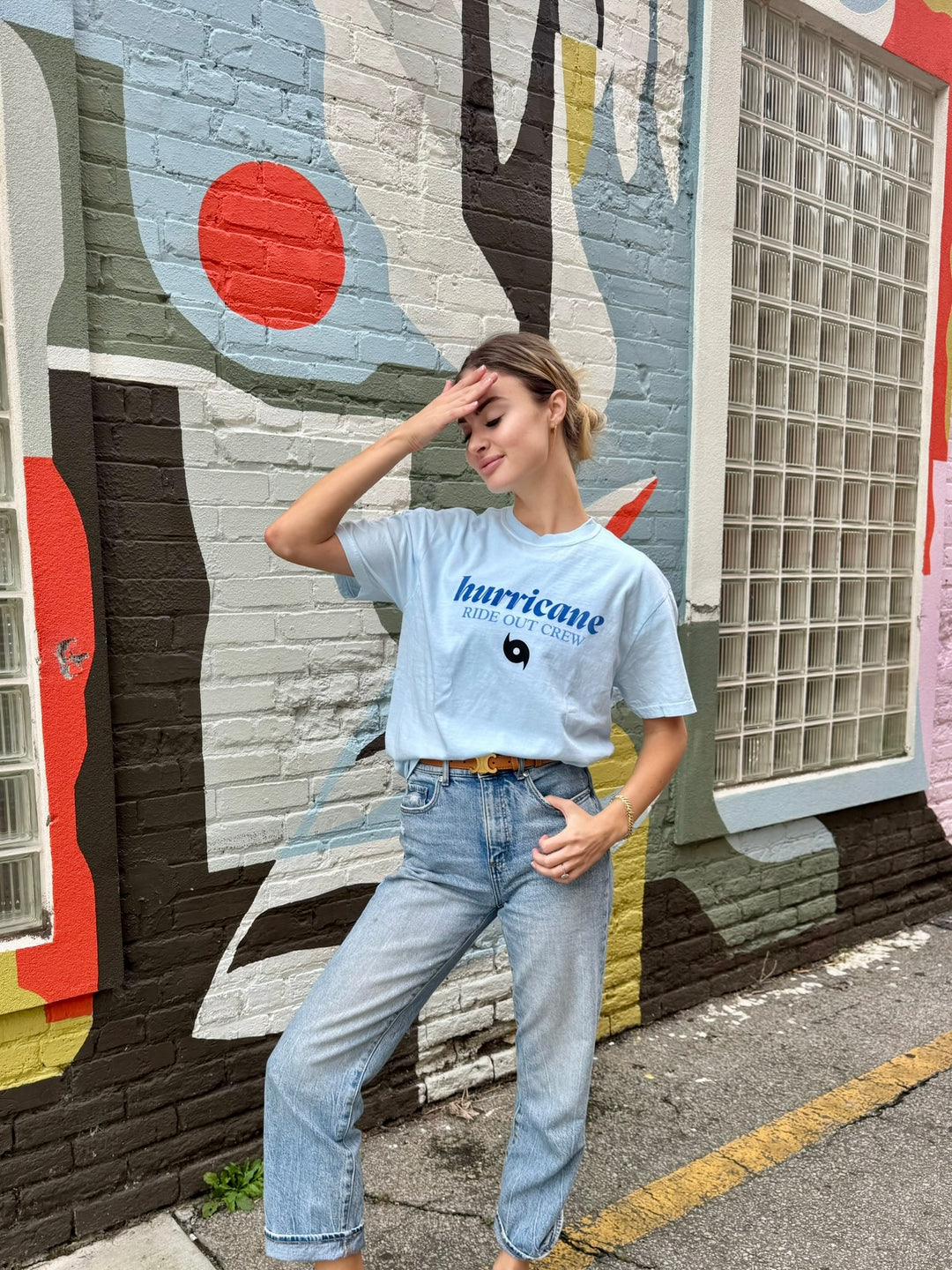Person wearing a light blue t-shirt with hurricane ride out crew text and blue jeans standing in front of a colorful mural.