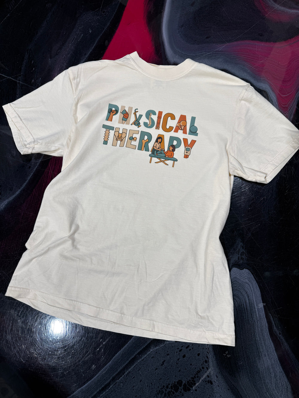 Flat lay of ivory t-shirt with 'Physical Therapy' text on a dark swirled surface
