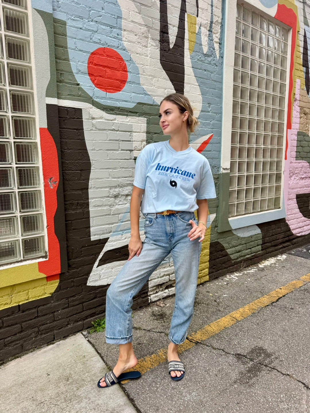 Woman wearing a light blue t-shirt with hurricane ride out crew text and jeans standing in front of a colorful mural.