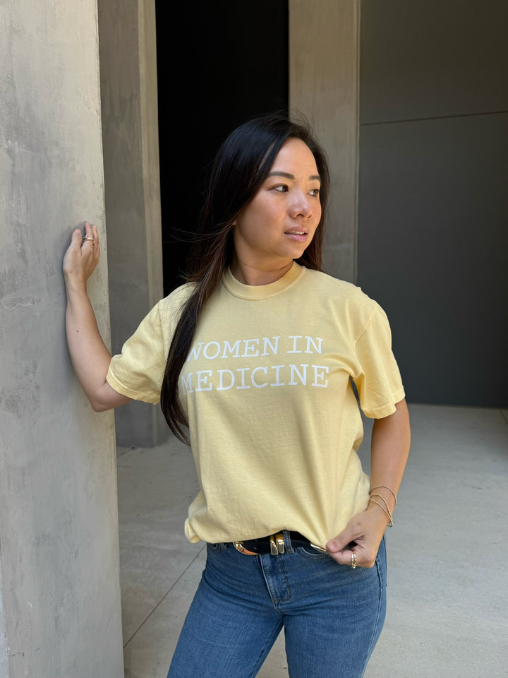 Person wearing a yellow 'Women in Medicine' t-shirt standing against a wall.