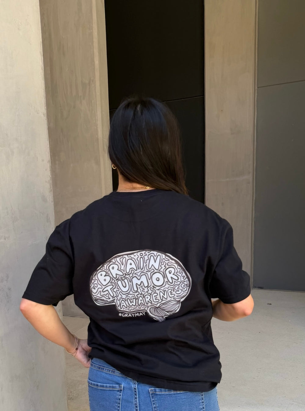 Person wearing a black t-shirt with a brain tumor awareness design on the back, standing against a plain wall.