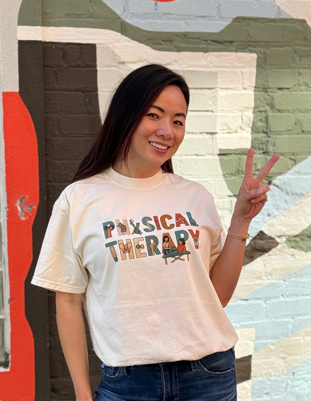 Woman wearing an ivory-colored 'Physical Therapy' t-shirt in front of a colorful mural.