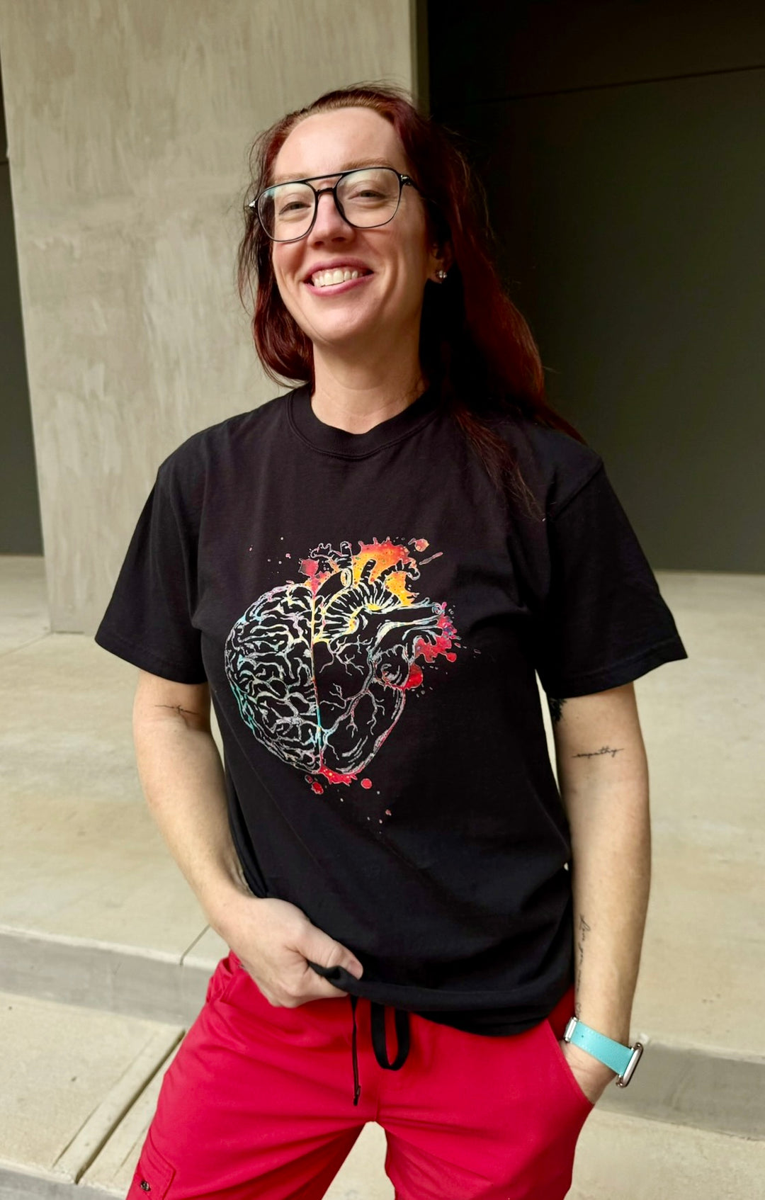 Ladyspinedoc wearing a black t-shirt with a colorful brain/heart graphic design, standing outdoors.