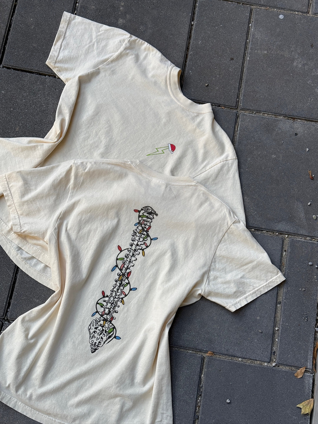 Front and back of ivory t-shirt with a green lightning bolt with Santa hat on front and spine decorated with Christmas lights on the back. 