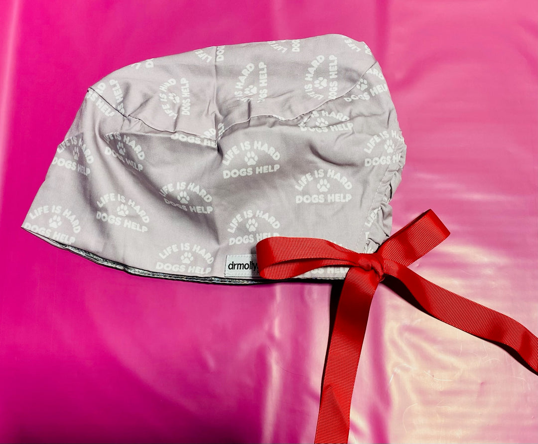 Gray scrub hat with white text and paw prints with a red ribbon on a pink background