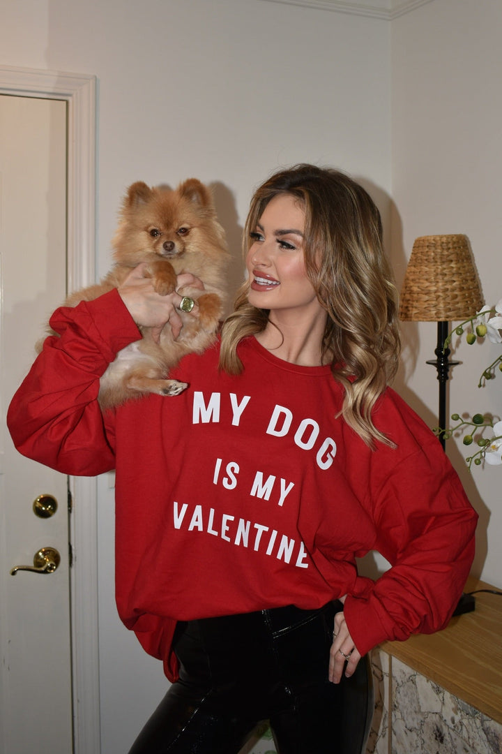 Woman wearing a red sweatshirt with 'My Dog is My Valentine' text, holding a small dog in a room.