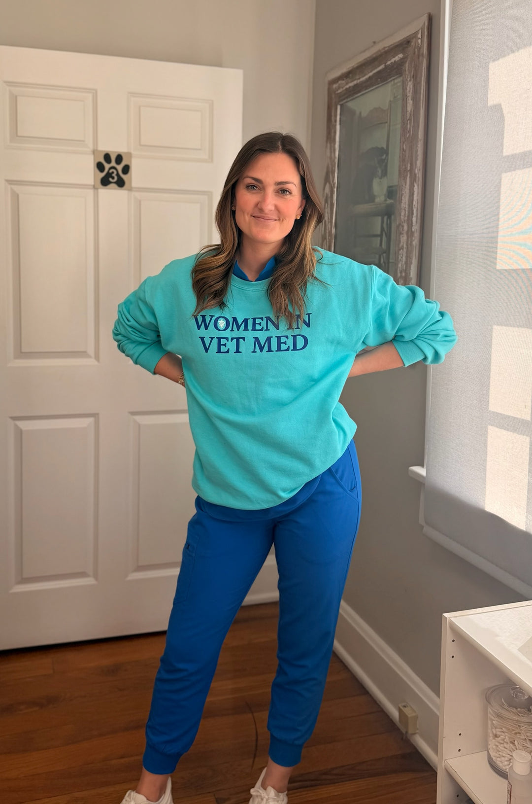 Dr. Molly Brinkmann wearing a teal sweatshirt with 'Women in Vet Med' text in a vet office setting.