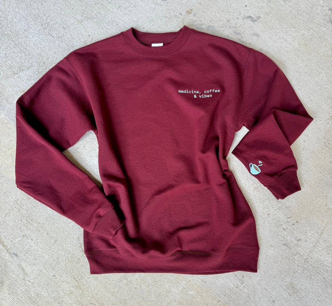 Maroon medicine, coffee & vibes sweatshirt with coffee cup on sleeve on a light gray surface
