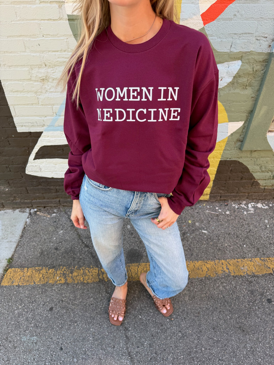 Person wearing a maroon sweatshirt with 'WOMEN IN MEDICINE' white text, standing on a street.