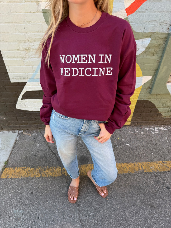Person wearing a maroon sweatshirt with 'WOMEN IN MEDICINE' white text, standing on a street.
