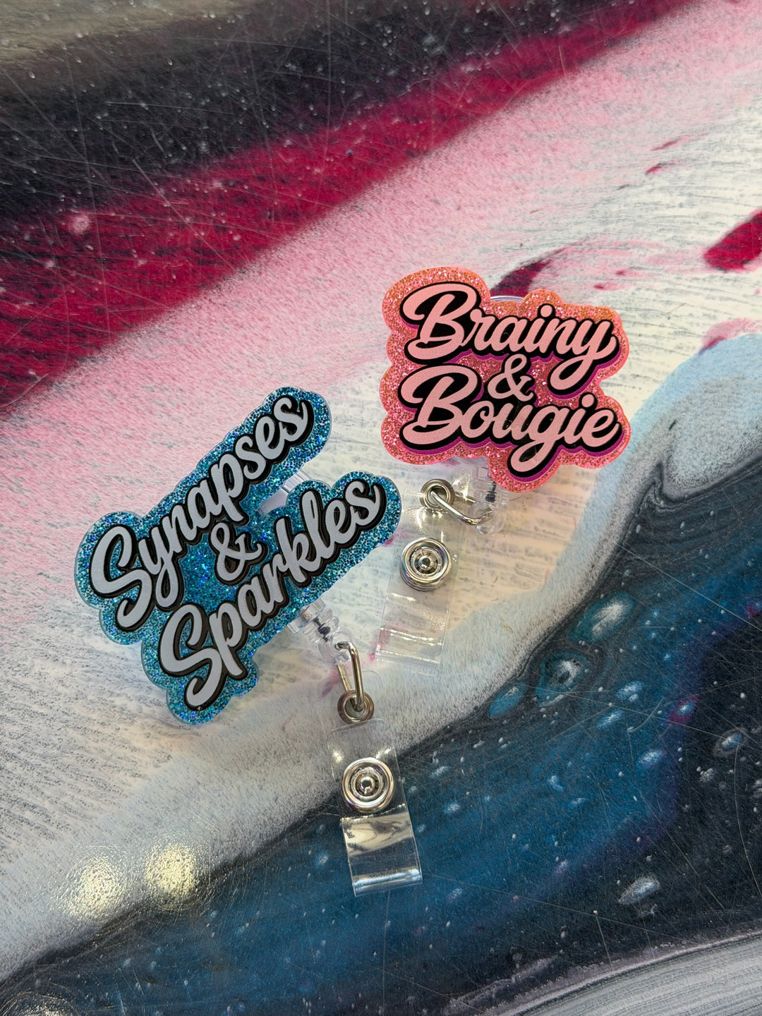 Two sparkly badge reels on a textured surface with 'Synapses & Sparkles' and 'Brainy & Bougie' text.