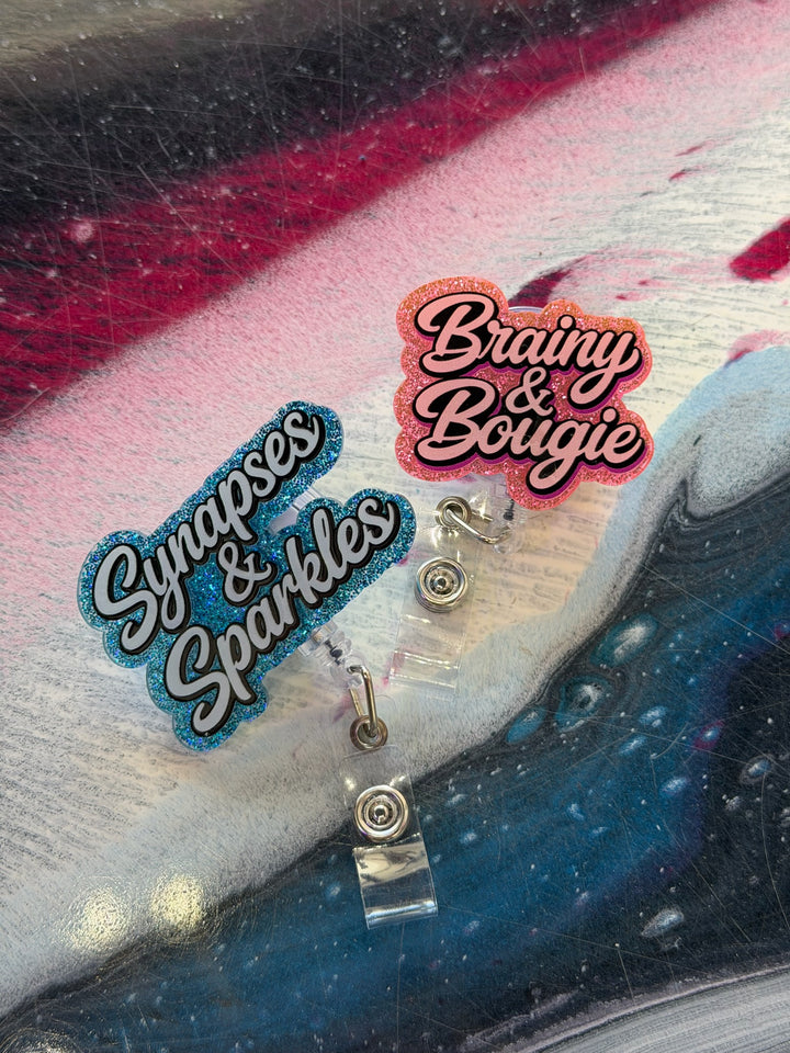 Two sparkly badge reels on a textured surface with 'Synapses & Sparkles' and 'Brainy & Bougie' text.