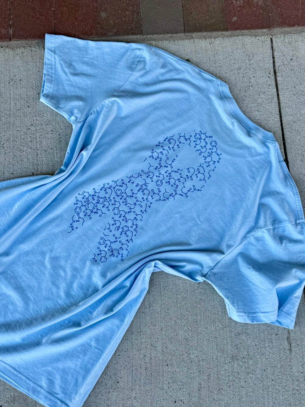 Back flat lay of a light blue diabetes awareness t-shirt with a ribbon design made up of insulin molecules on a concrete surface