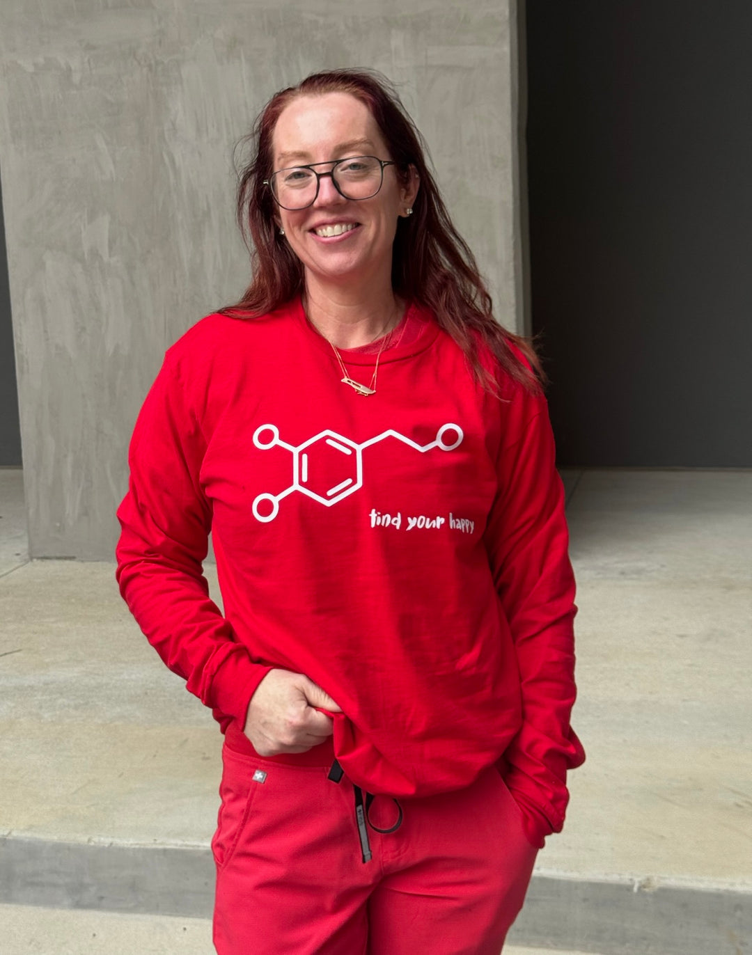 Ladyspinedoc wearing Dopamine Rush Long Sleeve Tee Shirt - Red with Dopamine Molecule Design and "Find Your Happy" Motto and standing against a concrete wall.