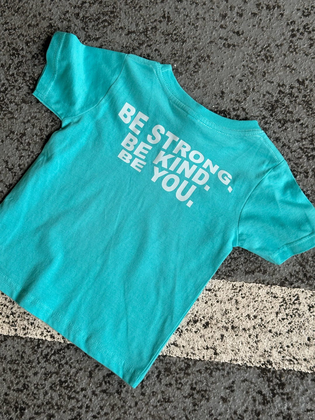 Back flat lay of light blue toddler t-shirt with “BE STRONG. BE KIND. BE YOU.” on the back in white lettering and displayed on pavement. 