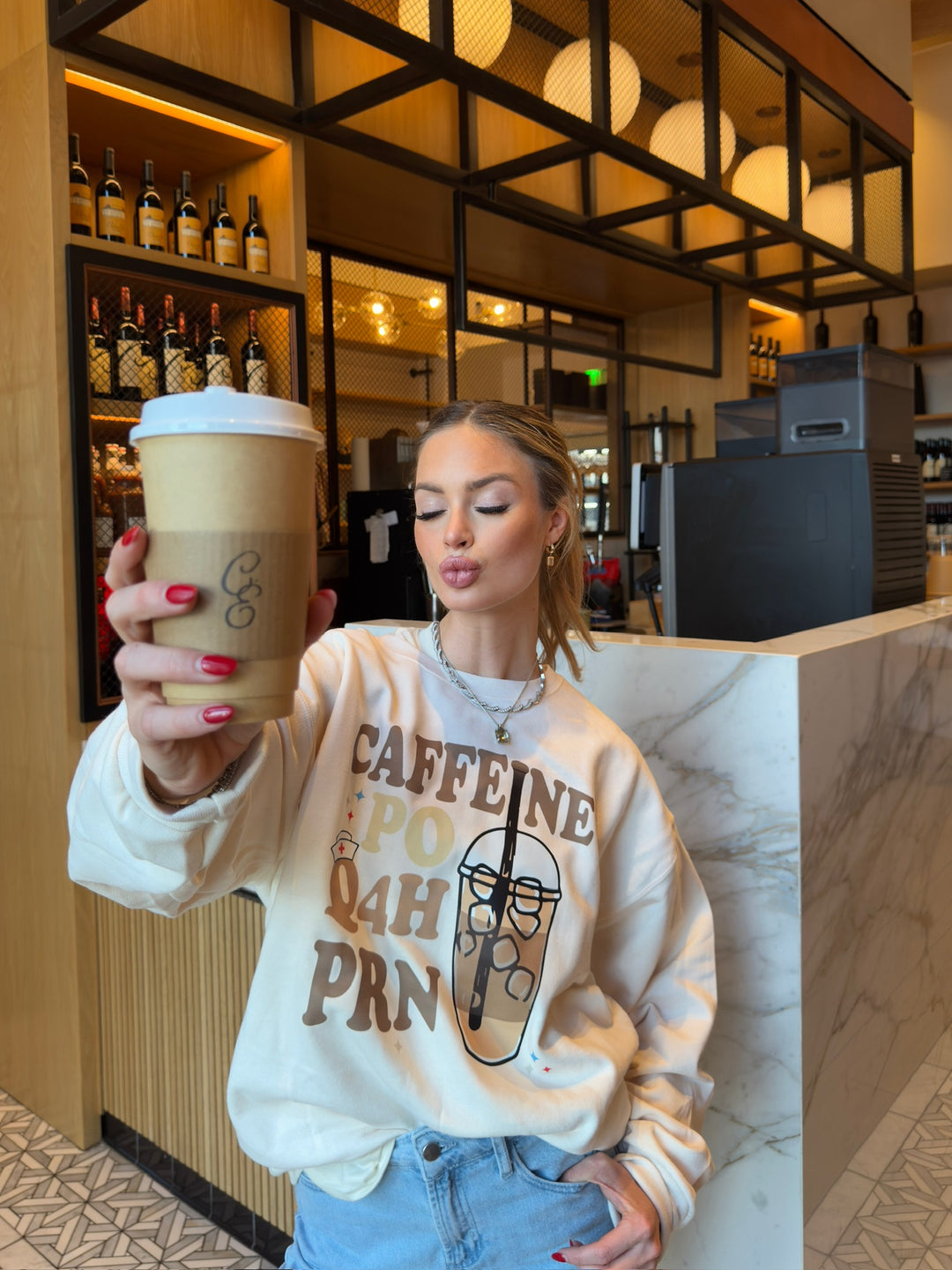Model pursing her lips and wearing Perk Up Rx Crewneck Sweatshirt while holding a coffee in a coffee shop
