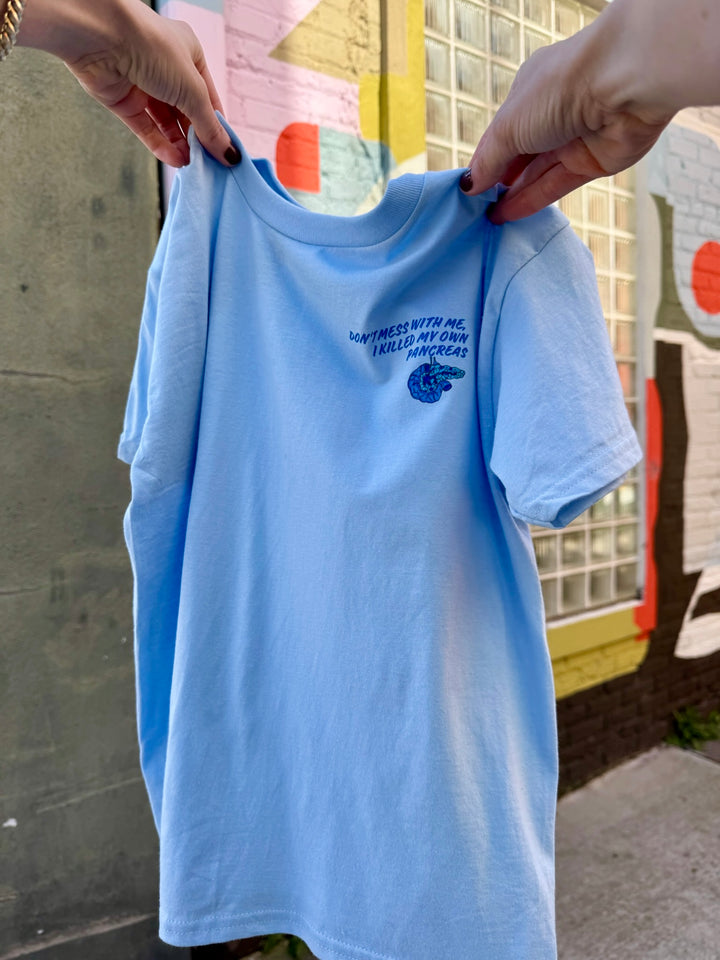 Light blue youth t-shirt with "Don't Mess With Me, I Killed My Own Pancreas" text held by hands against a colorful background