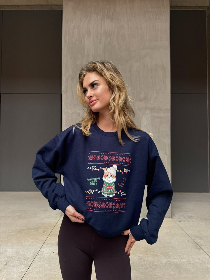 Woman wearing a navy blue sweatshirt with a festive cat design, standing against a plain wall.