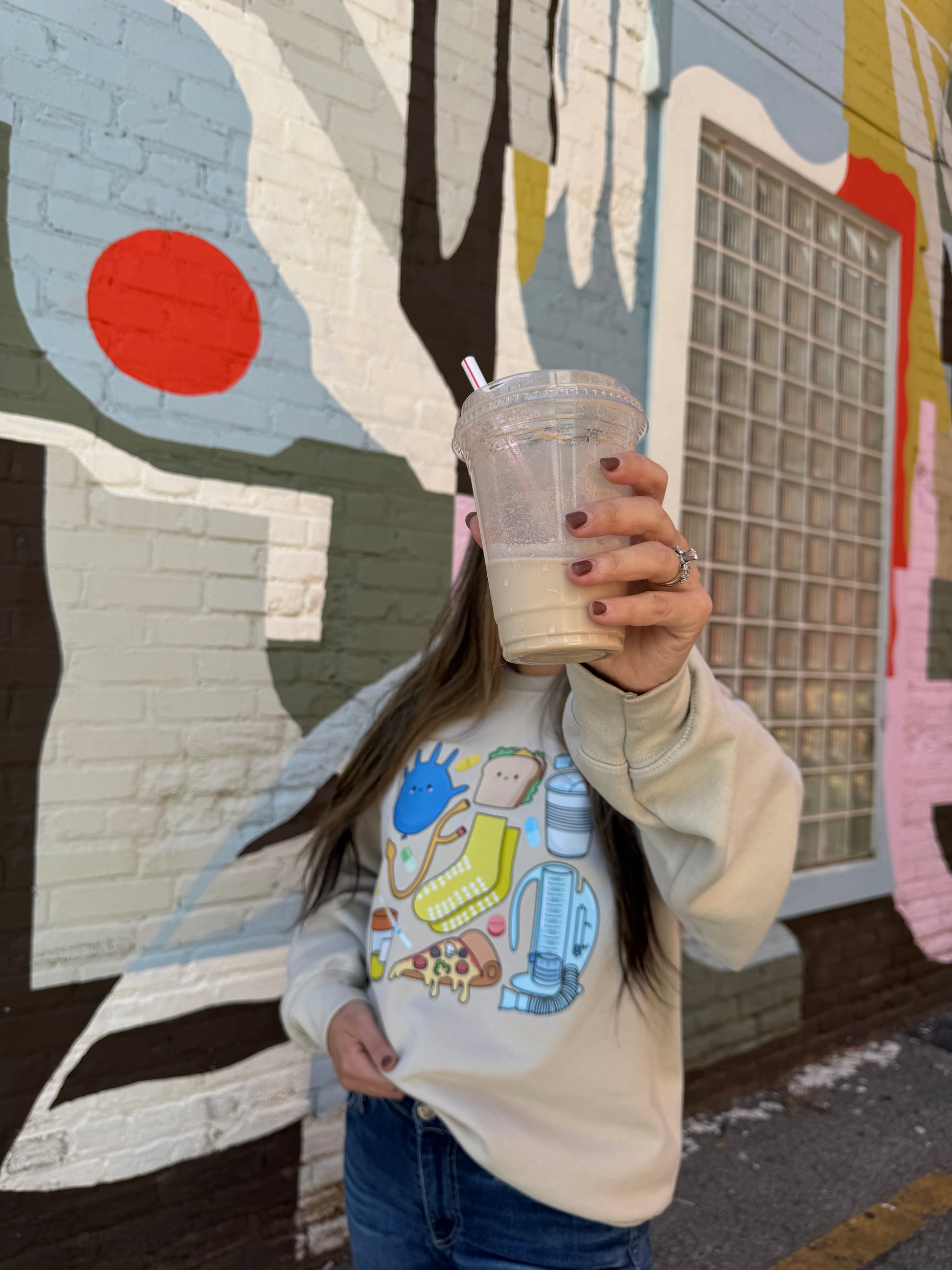 Women hiding her face with a cup and wearing a beige Nurse Sweatshirt with cartoon nurse memorabilia on the front. 