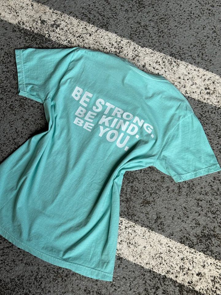 Back flat lay of BE YOU Tee Shirt – BE STRONG. BE KIND. BE YOU. in teal laying on pavement
