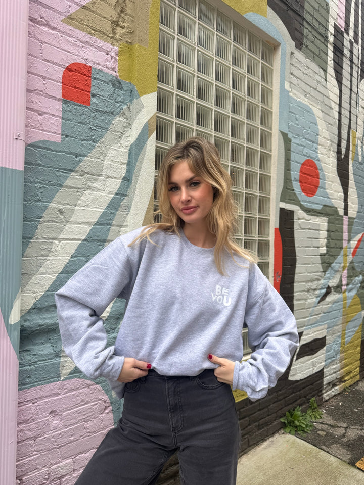Woman wearing a gray crewneck sweatshirt with “BE YOU.” printed on the front in bold white letters.