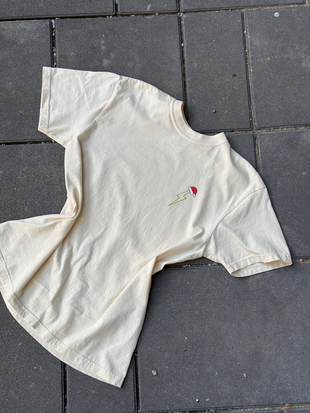 Ivory t-shirt with a green lightning bolt with Santa hat on top on upper left chest, laying on gray pavement