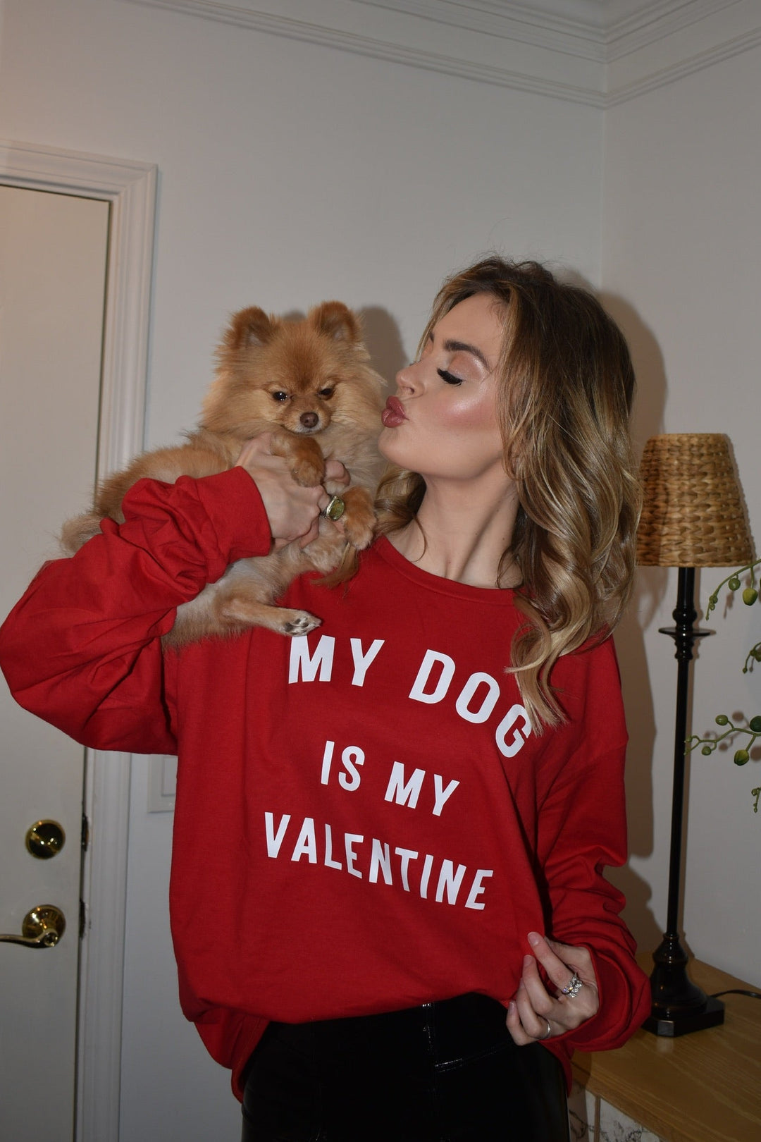Woman wearing a red sweatshirt with 'My Dog is my Valentine' text, holding a small dog indoors.