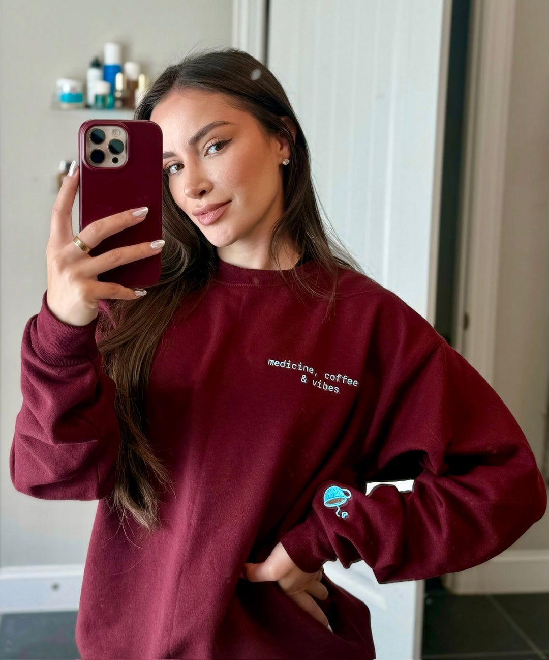 Dr Daisy wearing a maroon Medicine, Coffee & Vibes sweatshirt with coffee cup graphic on sleeve, taking a mirror selfie.